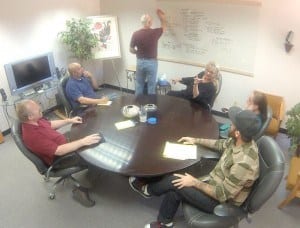 A group sitting around a round table with a whiteboard with writing.